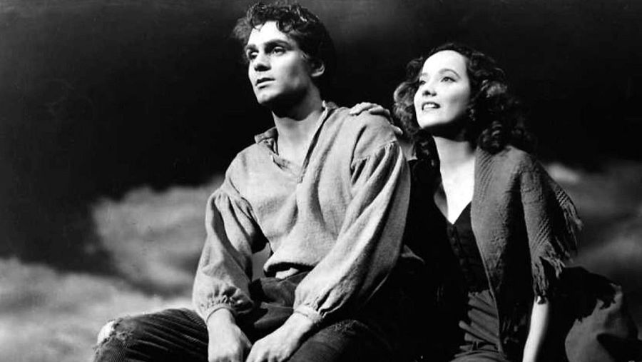 Una escena dramática en blanco y negro muestra a un hombre joven sentado con una expresión seria, junto a una mujer que lo toca con la mano, mirando hacia arriba con anhelo, con un cielo nublado de fondo.