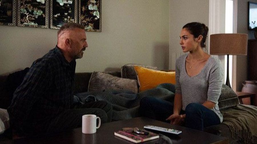 Kevin Costner y Gal Gadot comparten un momento en un sofá, posiblemente en una sala de estar. Se observa una mesa de centro con objetos como una taza, un cuaderno y un mando a distancia.