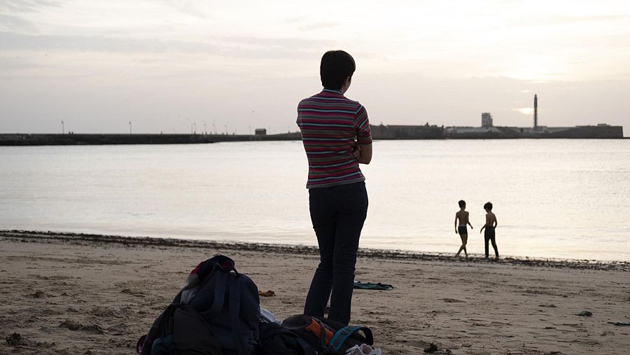 Una persona con camiseta a rayas y pantalones oscuros contempla el mar con los brazos cruzados en una playa al atardecer, mientras dos niños caminan por la orilla y mochilas yacen en la arena.
