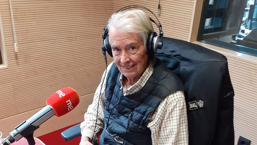 Manolo Velasco, con auriculares y gafas, está en un estudio de radio frente a un micrófono con el logo de RNE, vestido con camisa de cuadros y chaleco.