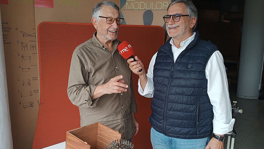 Dos hombres, uno con camisa caqui y otro con camisa blanca y chaleco, se encuentran en un interior. Uno de ellos gesticula mientras el otro sostiene un micrófono rojo, posiblemente en una entrevista. En la mesa hay una caja de madera y un objeto metálico.