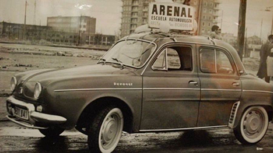 Un coche Renault antiguo, con la matrícula M-201141 y un letrero que indica "ARENAL ESCUELA AUTOMOVILISTA", se encuentra en una calle con edificios y personas caminando.