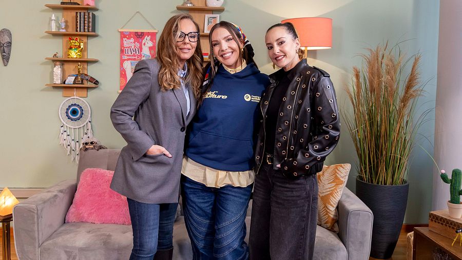 Tres mujeres posan en un estudio: una con gafas y camisa azul, otra con sudadera y pañuelo, y la tercera con chaqueta de cuero. El fondo muestra un sofá, una pared verde con estanterías y objetos decorativos.