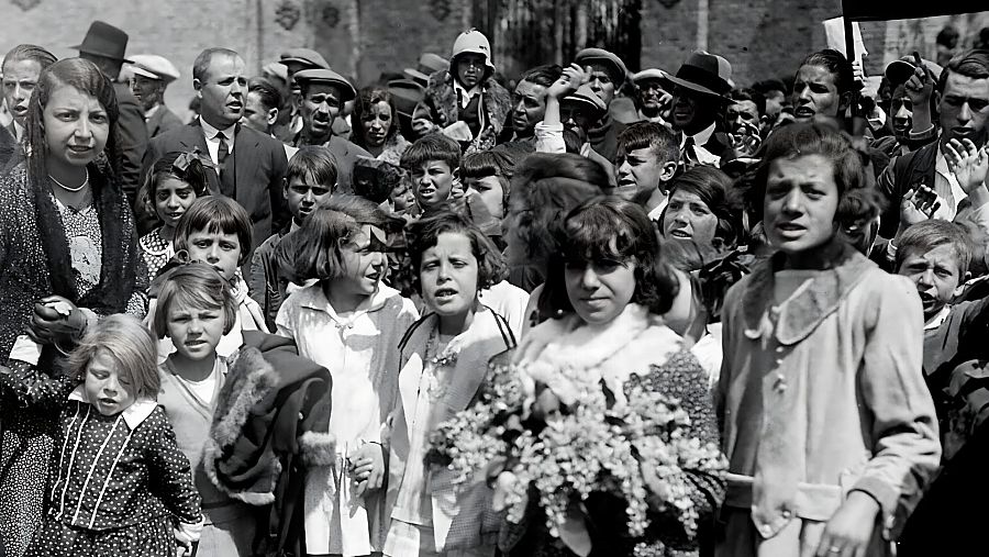 En una imagen en blanco y negro, un grupo numeroso de personas, entre ellas niños, se congrega en un cementerio. Algunos individuos portan flores y la mayoría observa atentamente, creando una atmósfera de solemnidad.