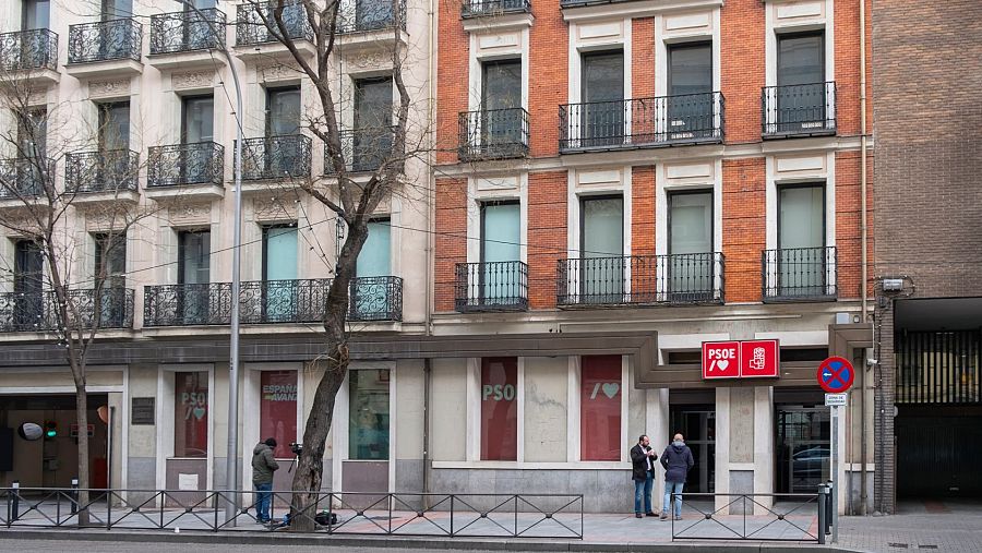 Fachada de dos edificios, uno claro y otro de ladrillo, con balcones y el logo del PSOE visible. Se aprecian carteles con las palabras "PSOE" y "España Avanza", junto a tres personas en la entrada.