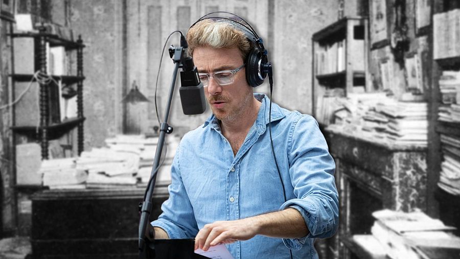 Víctor Clavijo, con gafas y auriculares, graba un documental frente a un micrófono, vestido con una camisa azul clara. El fondo es una imagen en blanco y negro de una habitación con libros.