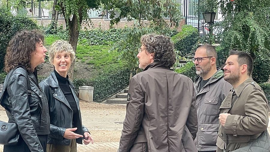 Un grupo de cinco personas posa al aire libre, con chaquetas de diferentes colores. Se distinguen dos mujeres con chaquetas de cuero negro y tres hombres, uno de ellos de espaldas, con chaquetas marrón, gris y verde oliva. El fondo muestra vegetación y una construcción de ladrillo.