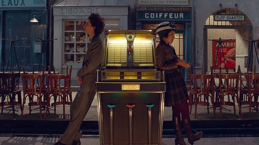 Una escena de cine muestra una jukebox antigua en una calle francesa, con un hombre y una mujer apoyados en ella. Se observan tiendas con letreros y mesas apiladas, creando una atmósfera nostálgica.