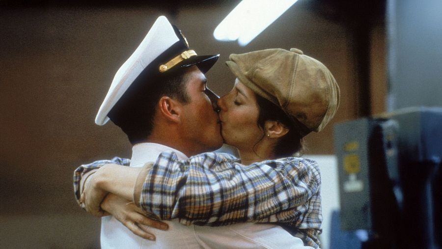 Un oficial de marina con uniforme blanco y gorra dorada comparte un beso apasionado con una mujer que lleva una camisa de cuadros y una gorra marrón, en un ambiente difuminado.