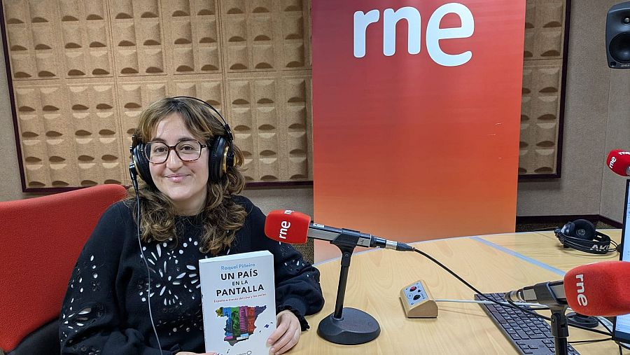 En un estudio de radio, una mujer con gafas y auriculares, vestida con un jersey negro, sostiene un libro. Frente a ella, se encuentran micrófonos y equipos de audio, con el logo de la emisora al fondo.