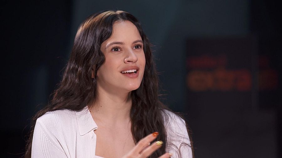 Rosalía, con cabello castaño ondulado y camisa blanca, posa para una entrevista. Sus uñas están pintadas con diseños variados, y el logo de la emisora se distingue en el fondo.