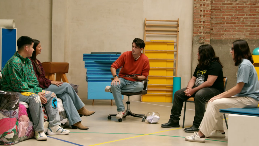 En un gimnasio, un hombre con jersey rojo y tablet conversa con cuatro adolescentes. Los jóvenes, vestidos de forma casual, escuchan atentamente mientras están sentados en círculo. Se observa equipamiento deportivo en el fondo.
