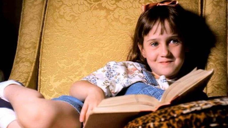 Una niña con peto vaquero y lazo rosa, presumiblemente Matilda, lee un libro en un sillón amarillo con estampado floral, apoyado sobre un cojín con estampado de leopardo, sonriendo levemente.