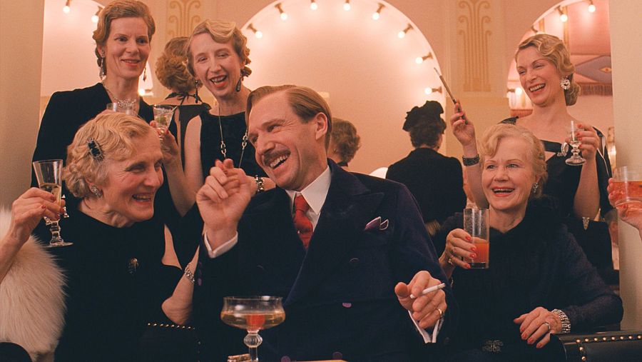 Ralph Fiennes, con traje oscuro y corbata roja, sonríe y gesticula mientras sostiene un cigarrillo, rodeado de mujeres en una celebración formal con copas y luces tenues.