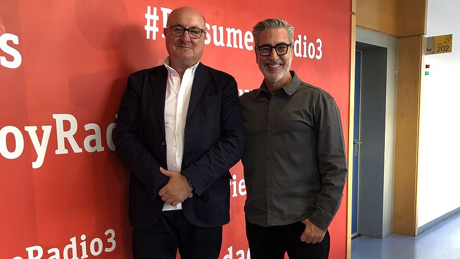 Dos hombres sonríen frente a una pared roja con un logo y un hashtag. Uno de ellos lleva gafas, traje oscuro y camisa blanca, mientras que el otro también lleva gafas y una camisa gris.