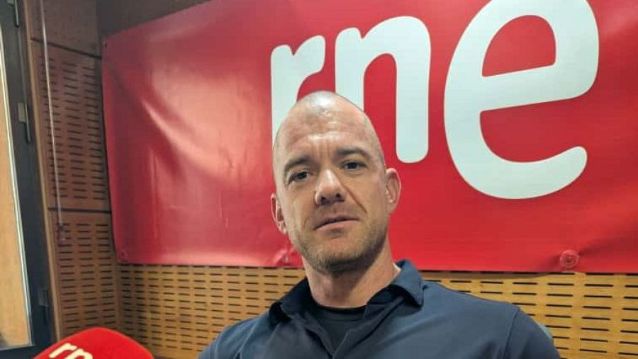Un hombre con barba y calvo, con una camisa oscura, se encuentra frente a un fondo rojo con un logo blanco. Un micrófono rojo con un logo similar se sitúa en la parte inferior izquierda.