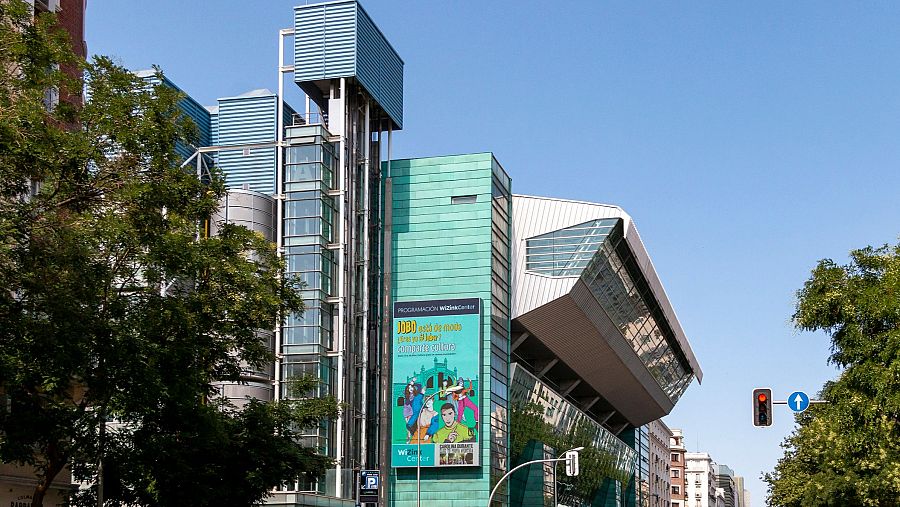 El edificio de arquitectura moderna, con elementos de cristal y metal, exhibe un cartel publicitario que anuncia un evento con personajes de dibujos animados. En primer plano, árboles y un semáforo con la luz roja encendida.