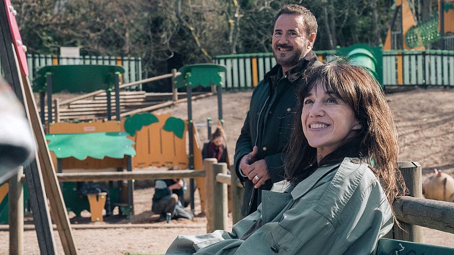 En un parque infantil, Charlotte Gainsbourg y José Garcia posan sonrientes. Ella está sentada en un banco con una gabardina verde oliva, mientras él, de pie, viste una chaqueta oscura.