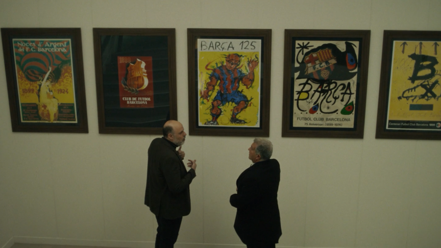 Dos hombres, uno con barba y calvicie y otro con cabello canoso, conversan frente a una pared donde se exhiben varios carteles enmarcados. Los carteles parecen estar relacionados con un club deportivo, mostrando escudos y diseños conmemorativos.