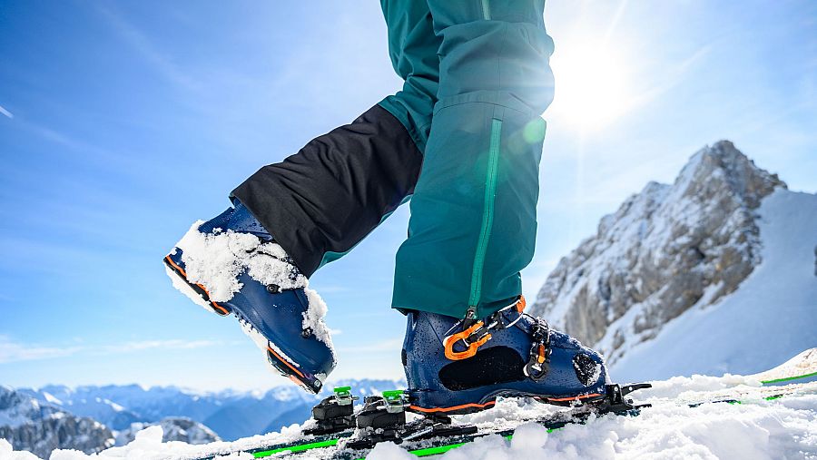 Una persona con pantalones de esquí verde azulado y botas azules esquía en la nieve, con esquíes verdes y un paisaje montañoso como fondo.