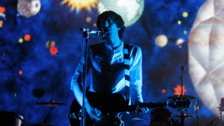 Un guitarrista de Snow Patrol, con camisa blanca y correa a juego, toca en un escenario iluminado en azul, con una pantalla LED mostrando imágenes abstractas de fondo.