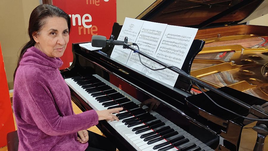 Una pianista toca un piano de cola negro, con partituras y un micrófono sobre el instrumento. Se aprecia el logo de una emisora en la esquina superior izquierda.