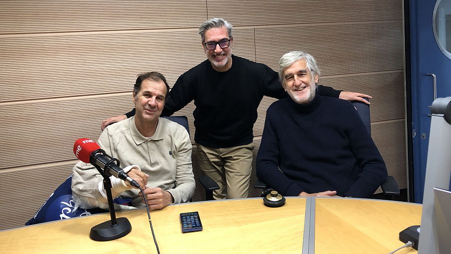 Tres hombres sonríen en un estudio de radio. Uno está sentado frente a un micrófono, otro de pie y el tercero sentado a la derecha. En la mesa hay un teléfono móvil.