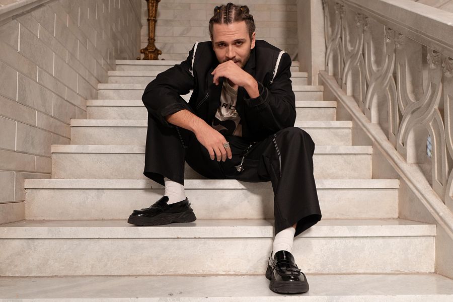 En la imagen, un hombre con trenzas y un anillo en la mano izquierda, posa sentado en unas escaleras de mármol, vistiendo una chaqueta negra con detalles blancos, camiseta estampada, pantalones anchos y zapatos tipo mocasín.
