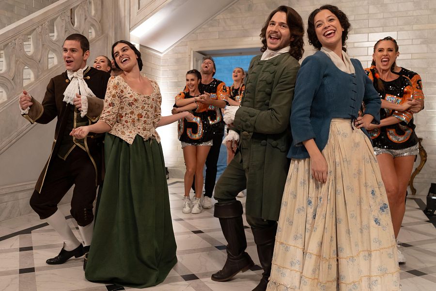Un grupo diverso posa en un espacio con una escalera de mármol. Se distinguen personas con trajes de época y otras con ropa moderna, como camisetas con el número 55, todos sonriendo.