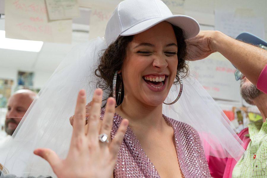 Elena Navarro, caracterizada, sonríe y muestra un anillo, llevando un top rosa brillante y una gorra con velo. Se encuentra en un área de preparación con otras personas.