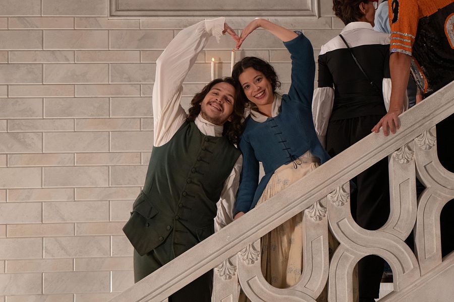 Los actores Marcos Orengo y Elena Navarro, caracterizados, sonríen y forman un corazón con sus manos en unas escaleras de piedra. Él viste camisa y chaleco, ella blusa y falda larga.