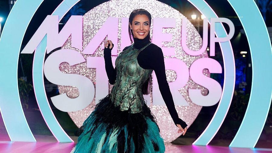 Una mujer con un top negro y un corsé verde posa frente a un letrero que dice "Make Up Stars" en letras rosas y blancas, con arcos azules de fondo, sobre un suelo rosa brillante.