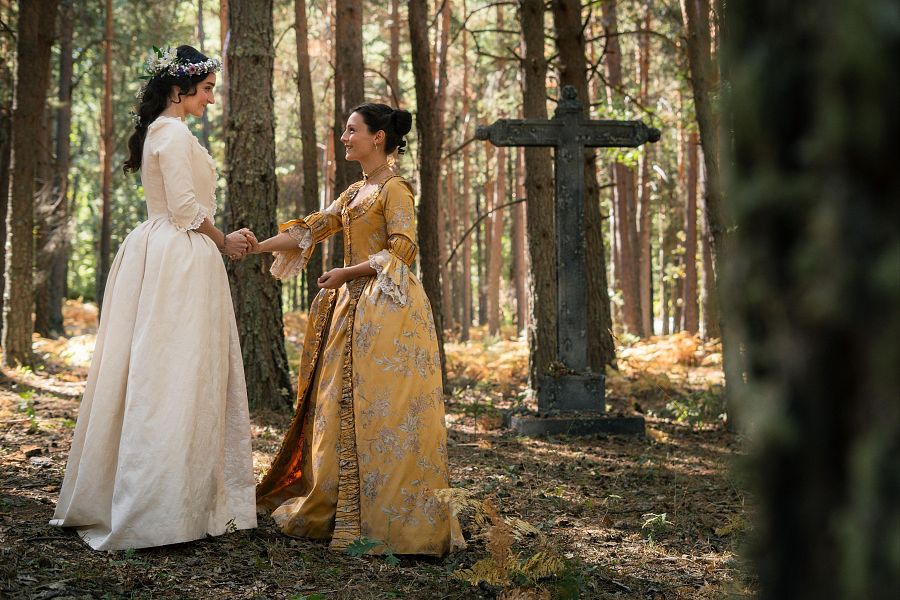 Dos mujeres, vestidas con trajes de época, se encuentran en un bosque. Una lleva un vestido blanco y una corona de flores, mientras que la otra viste un vestido amarillo, ambas se toman de la mano.