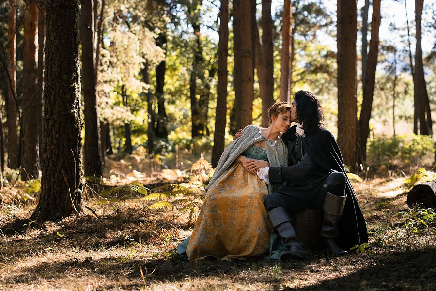 Una mujer con un vestido de época y un chal gris está sentada sobre un tronco en un bosque, mientras un hombre vestido de negro la besa en la frente.