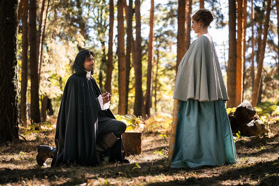 En un entorno boscoso, un hombre vestido con traje oscuro y capa se arrodilla frente a una mujer con un vestido largo y una capa, posiblemente para proponerle matrimonio.