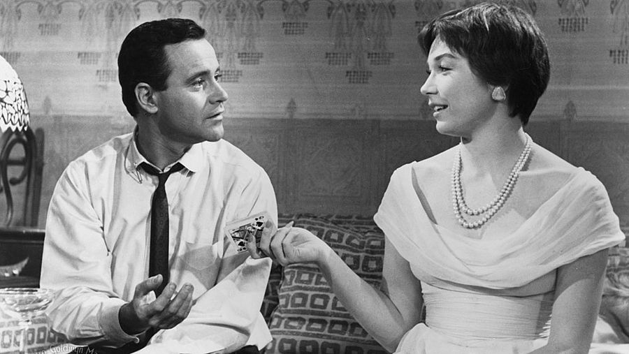 Una imagen en blanco y negro captura a un hombre y una mujer en una escena de una película. Él, con camisa y corbata, sostiene un papel; ella, con vestido y collar de perlas, le observa. El entorno se compone de un papel tapiz floral y un cojín con diseño.
