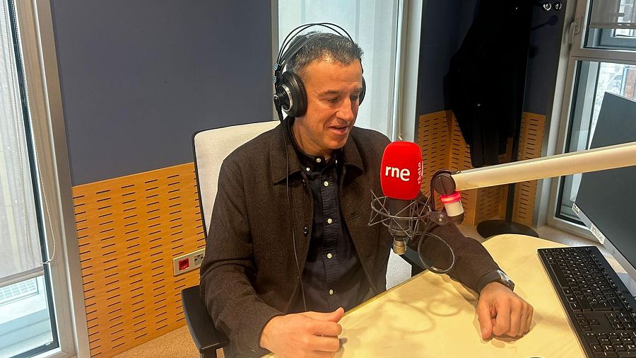 May Peus, con auriculares y camisa oscura, está sentado frente a un micrófono con el logo de una emisora, posiblemente en Barcelona, con un teclado negro en la mesa y una pared de madera al fondo.