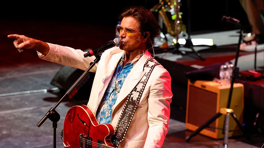 En un escenario iluminado, un hombre con gafas de sol y chaqueta blanca toca la guitarra roja, con su nombre en la correa, mientras canta frente a un micrófono. Se observan otros músicos y un amplificador.