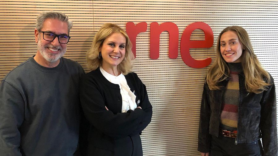 Tres personas posan frente a una pared con un logo rojo. Un hombre con gafas y barba, una mujer con blusa blanca y chaqueta negra, y otra mujer con chaqueta oscura y jersey a rayas.