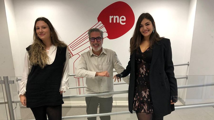 Tres individuos, incluyendo a una mujer con blusa y chaleco, un hombre con barba y gafas, y otra mujer con vestido, posan delante de un fondo blanco con un logotipo rojo.