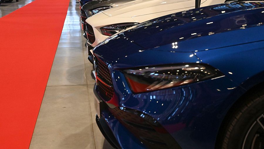 Una fila de coches de diferentes colores, incluyendo uno azul brillante con detalles cromados y parrilla negra, se exhiben sobre un suelo de baldosas grises, con una alfombra roja a un lado.