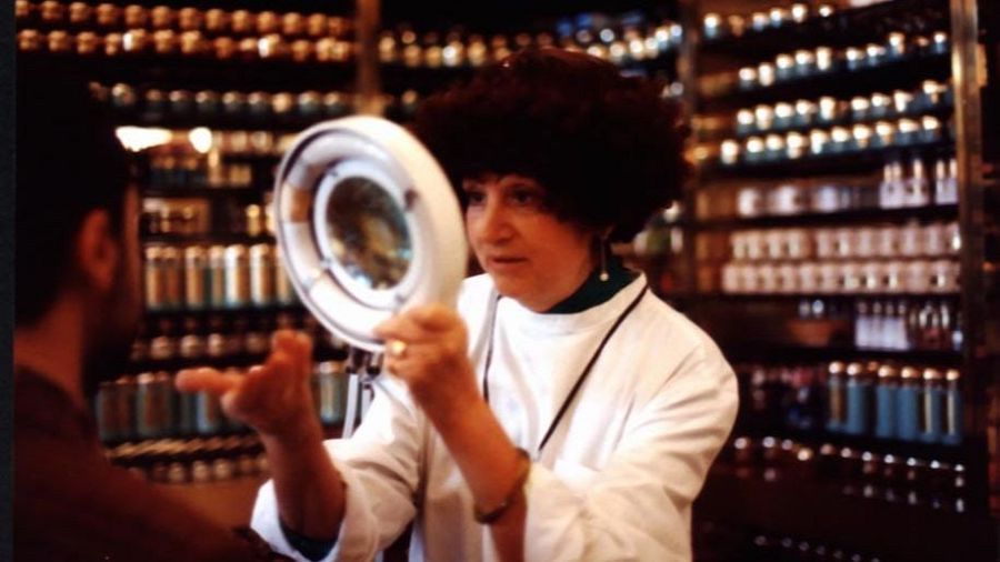 Paquita Ors, con un peinado afro, utiliza una lupa para examinar la piel de un cliente. Viste una bata blanca y lleva un collar y pendientes pequeños. El fondo está lleno de frascos y botellas.