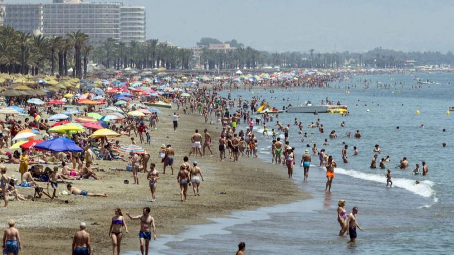 Una playa concurrida en un día soleado, donde la arena está llena de personas tomando el sol y disfrutando del agua. Se ven sombrillas de colores y edificios altos al fondo.