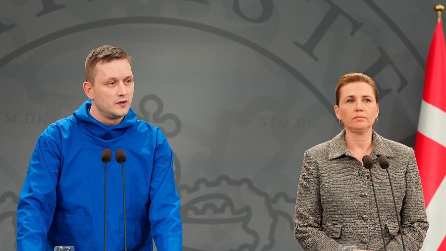 Dos figuras políticas, el presidente de Groenlandia y la primera ministra de Dinamarca, se encuentran en una rueda de prensa. Uno viste una sudadera azul y la otra una chaqueta gris, ambos frente a micrófonos con un fondo gris y la bandera danesa.