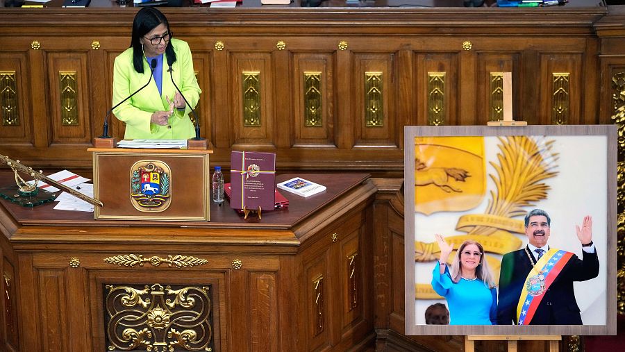 En la Asamblea Nacional de Caracas, una mujer vestida de verde lima se dirige a la audiencia desde un podio con el escudo nacional, mientras gesticula. A su lado, una fotografía enmarcada muestra a dos personas saludando.