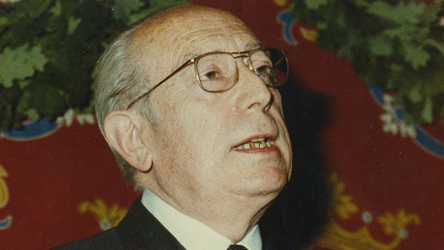 En primer plano, se observa a un hombre mayor con gafas y traje oscuro, posiblemente en un discurso. El fondo incluye un tapiz decorativo con detalles en rojo, dorado y azul, y una guirnalda.