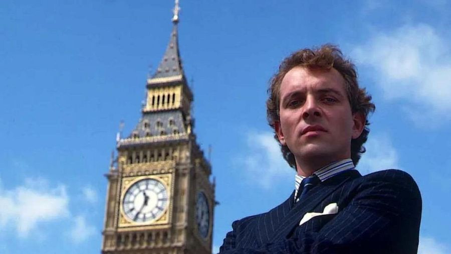 Un hombre con traje y corbata azules, de pie con los brazos cruzados, se encuentra frente a una torre de reloj gótica, con el cielo azul como telón de fondo.