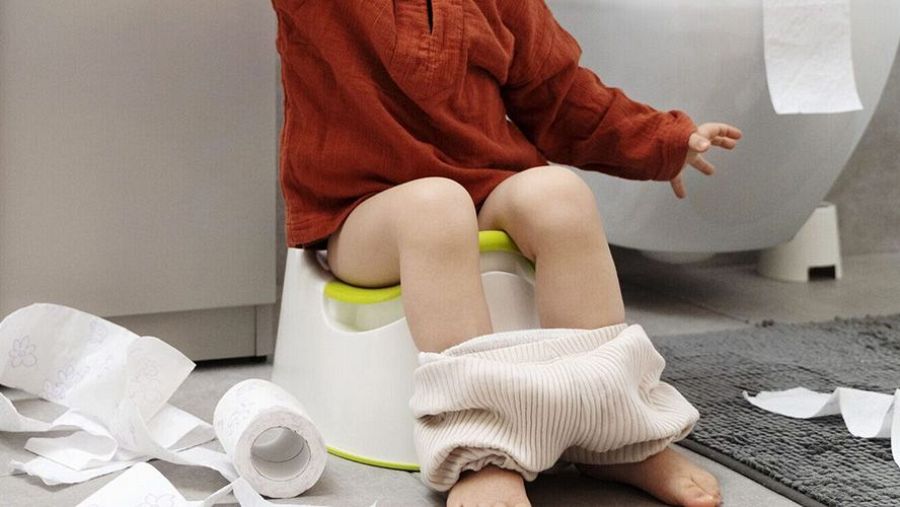 Un niño pequeño, con la ropa bajada, está sentado en un orinal blanco con borde verde, rodeado de papel higiénico. Se observa un rollo de papel casi vacío y un mueble de baño al fondo.