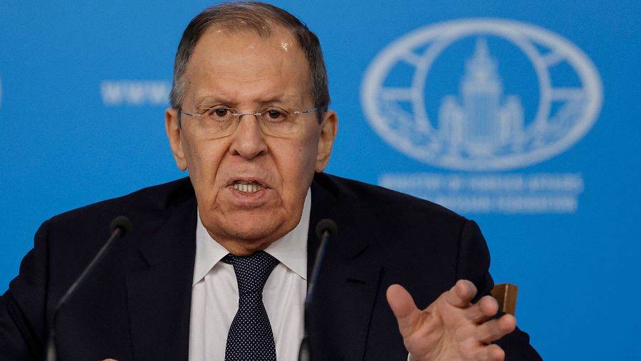Sergei Lavrov, con traje oscuro y corbata, habla en una conferencia de prensa, gesticulando con las manos. Detrás de él, un fondo azul con un emblema y micrófonos.