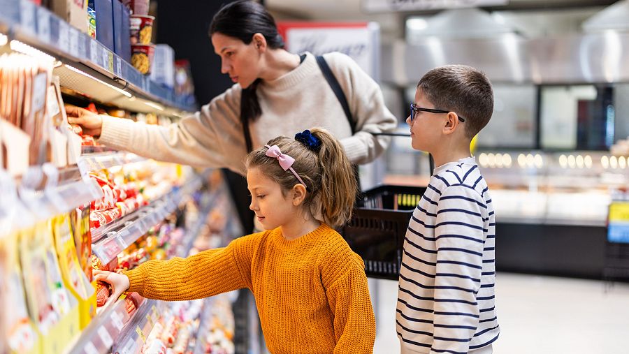 Una familia, compuesta por una mujer y dos niños, compra en un supermercado. La mujer, con el pelo recogido, alcanza un producto. Los niños observan los productos en los estantes, uno con un lazo rosa y otro con gafas.